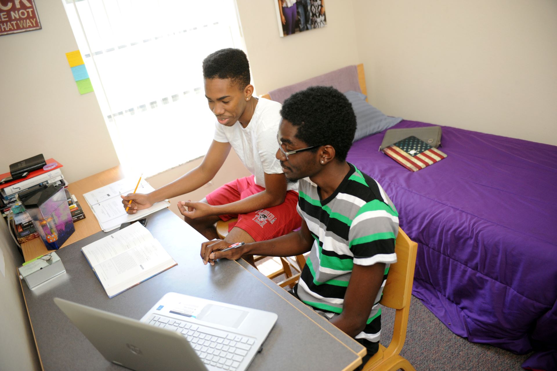 Two students studying together at a desk in a dorm room with a bed in the background